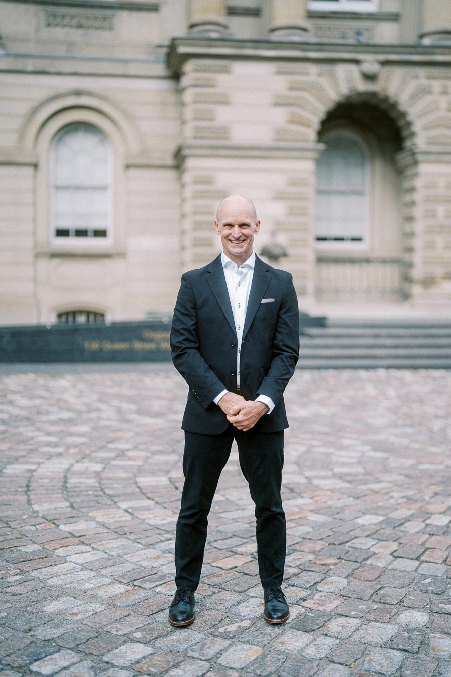 Man in a suit smiling confidently in front of a historic building with arched windows and cobblestone path.