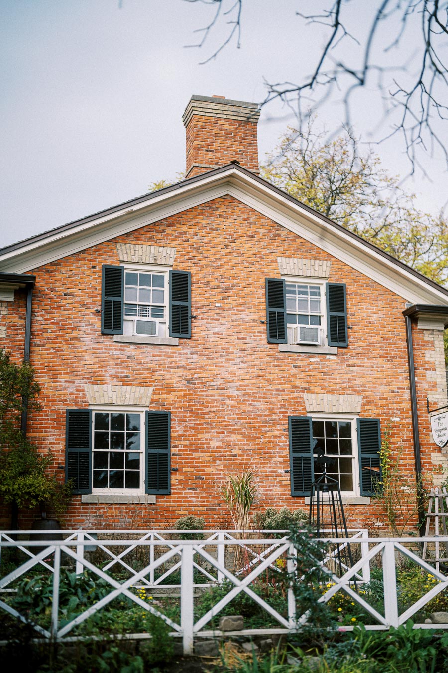 Historic red brick house with black shutters and a wooden fence surrounded by a garden, set against a cloudy sky.