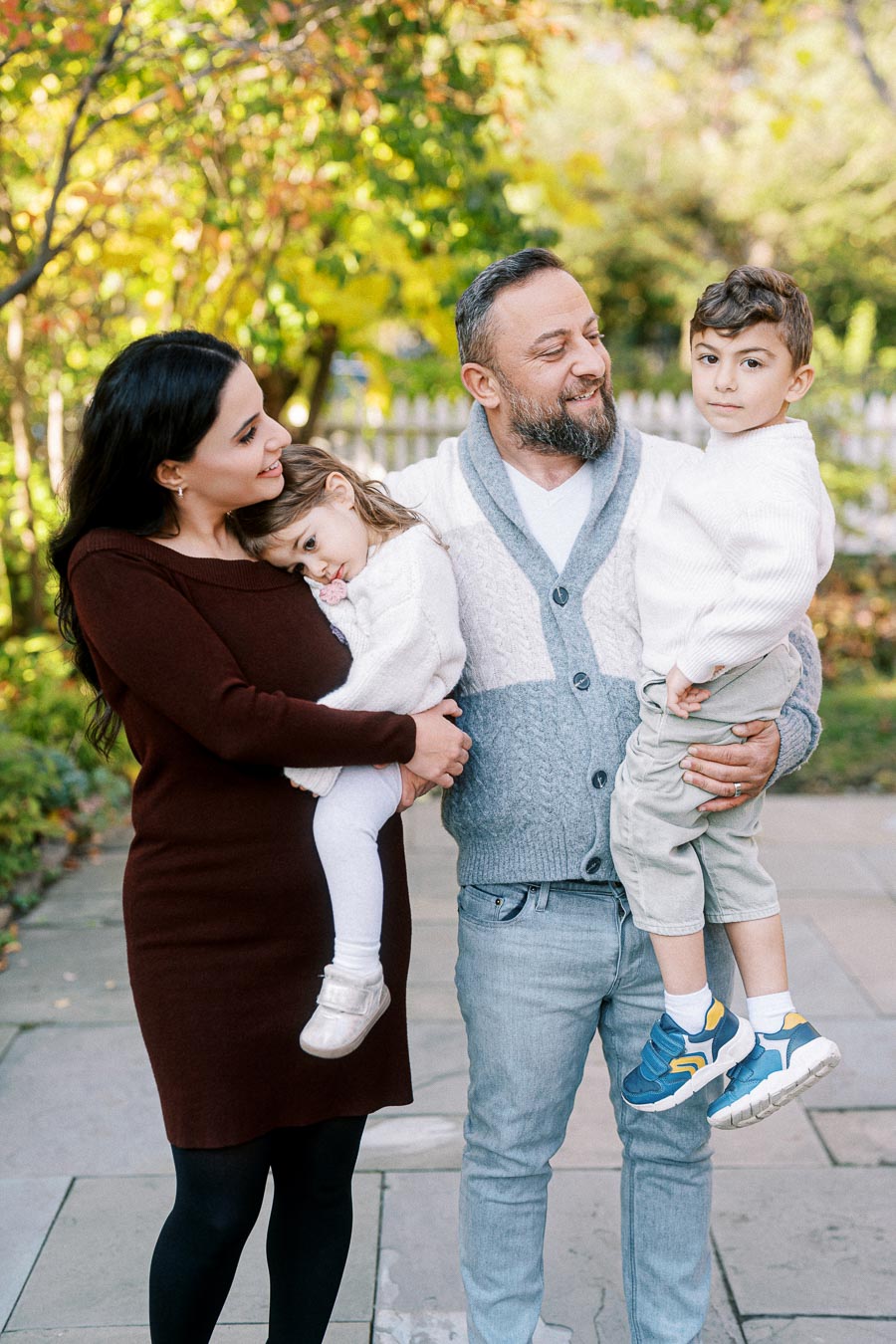 A happy family of four enjoying a sunny day in the park, with colorful autumn foliage in the background. Parents are smiling while holding their young children, who are dressed in cozy winter outfits. The scene captures warm family bonding in nature.