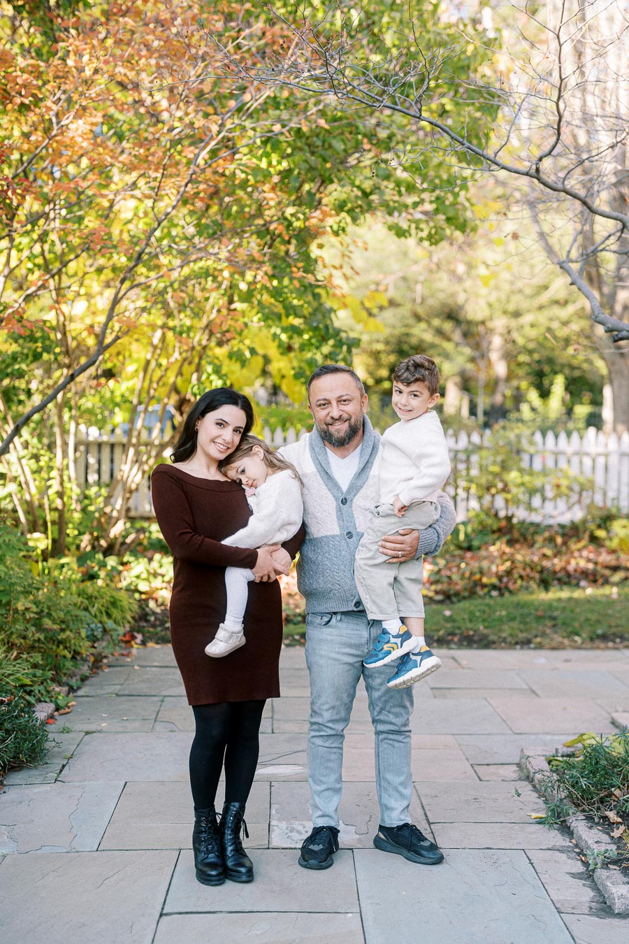 A family of four smiling outdoors in an autumn setting, with colorful leaves in the background.