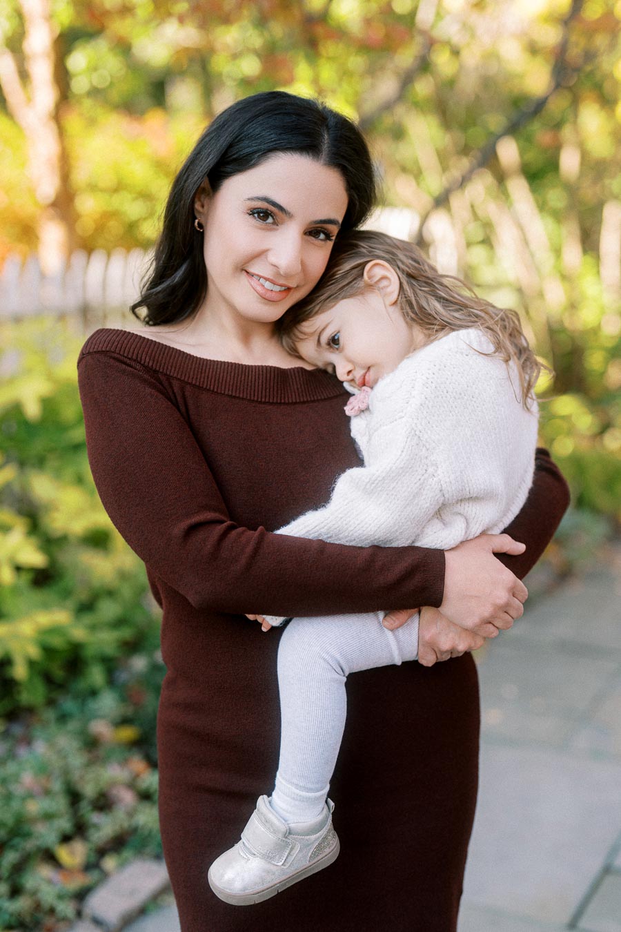 A mother in a brown sweater lovingly holds her daughter wearing a white sweater and leggings in a garden setting with soft autumnal hues.