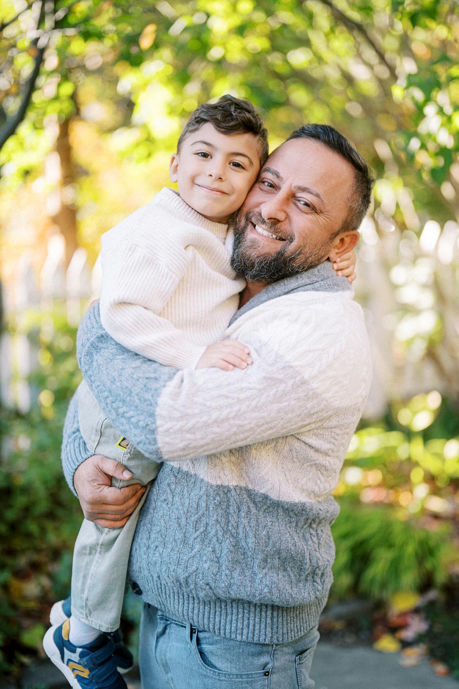 Father and son enjoying a happy moment in a green, sunlit garden, both wearing cozy sweaters.