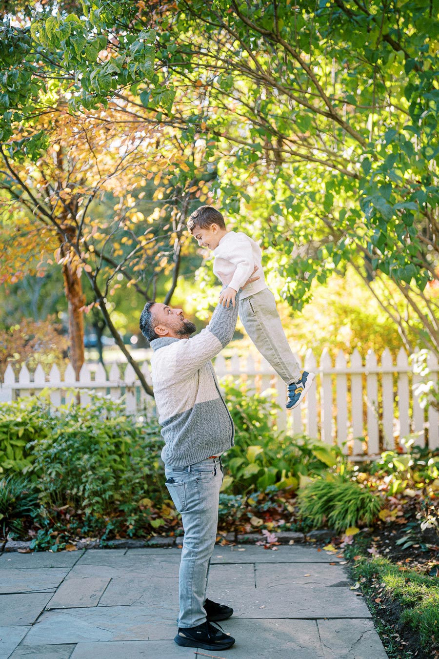 A father joyfully lifting his son in a sunny garden, surrounded by lush greenery and autumn foliage near a white picket fence.