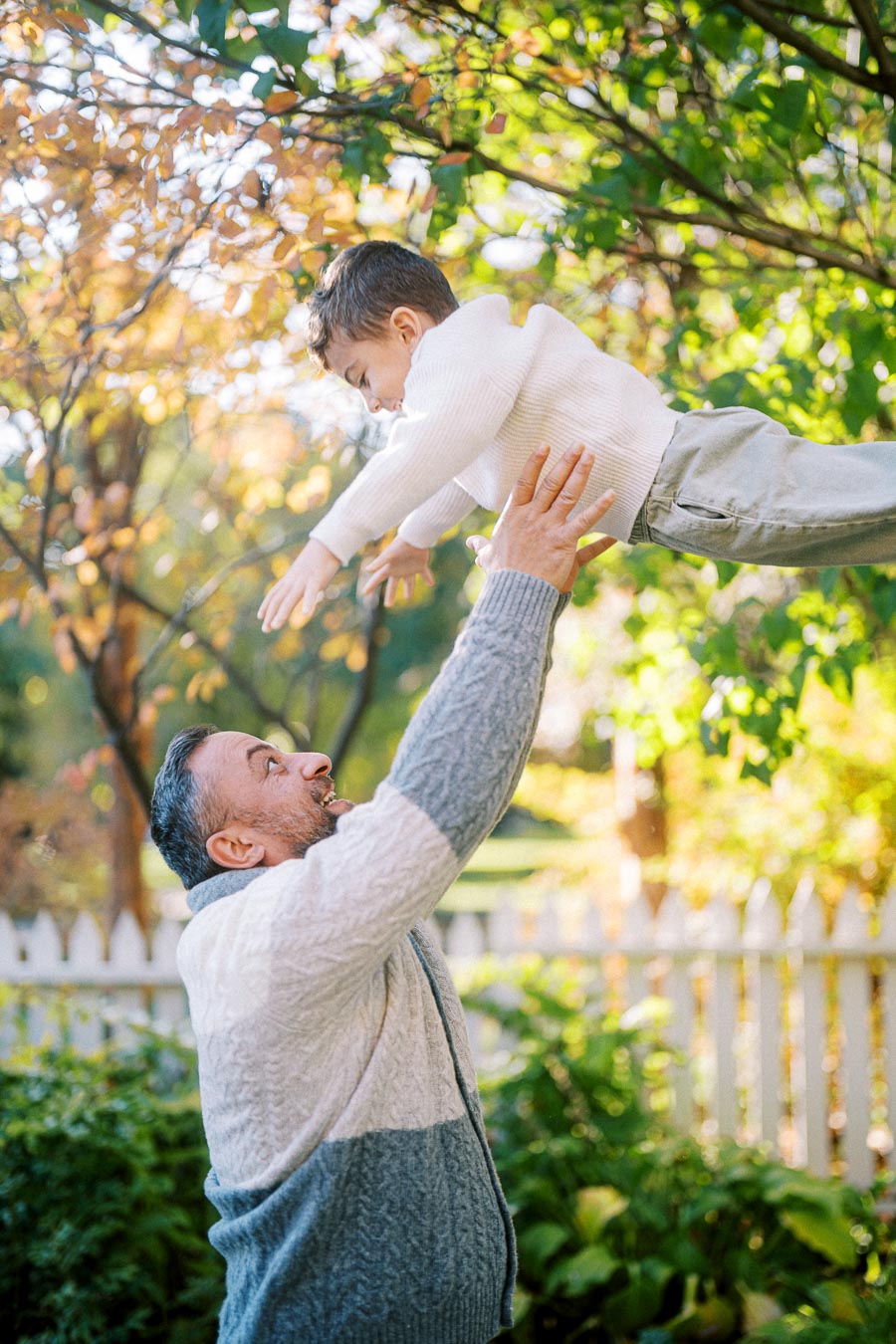 A man in a cozy sweater lifts a child in a fall garden, surrounded by colorful autumn leaves and a white picket fence.