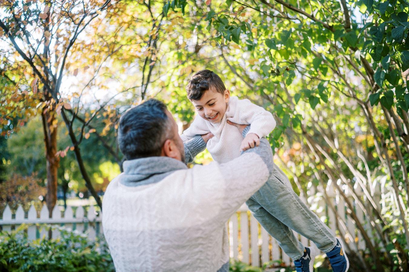 Father lifting and playing with his smiling son outdoors in a garden surrounded by lush green trees and a wooden fence, capturing a joyful family moment in nature.