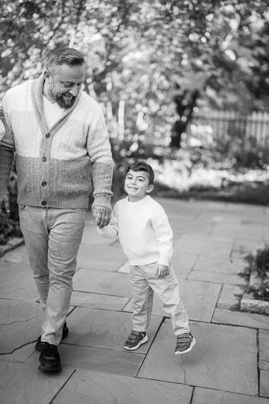 A bearded man and a young boy smiling and holding hands while walking outdoors on a stone path surrounded by lush greenery. The man is wearing a cozy sweater, and the boy is dressed in a light sweater and jeans.