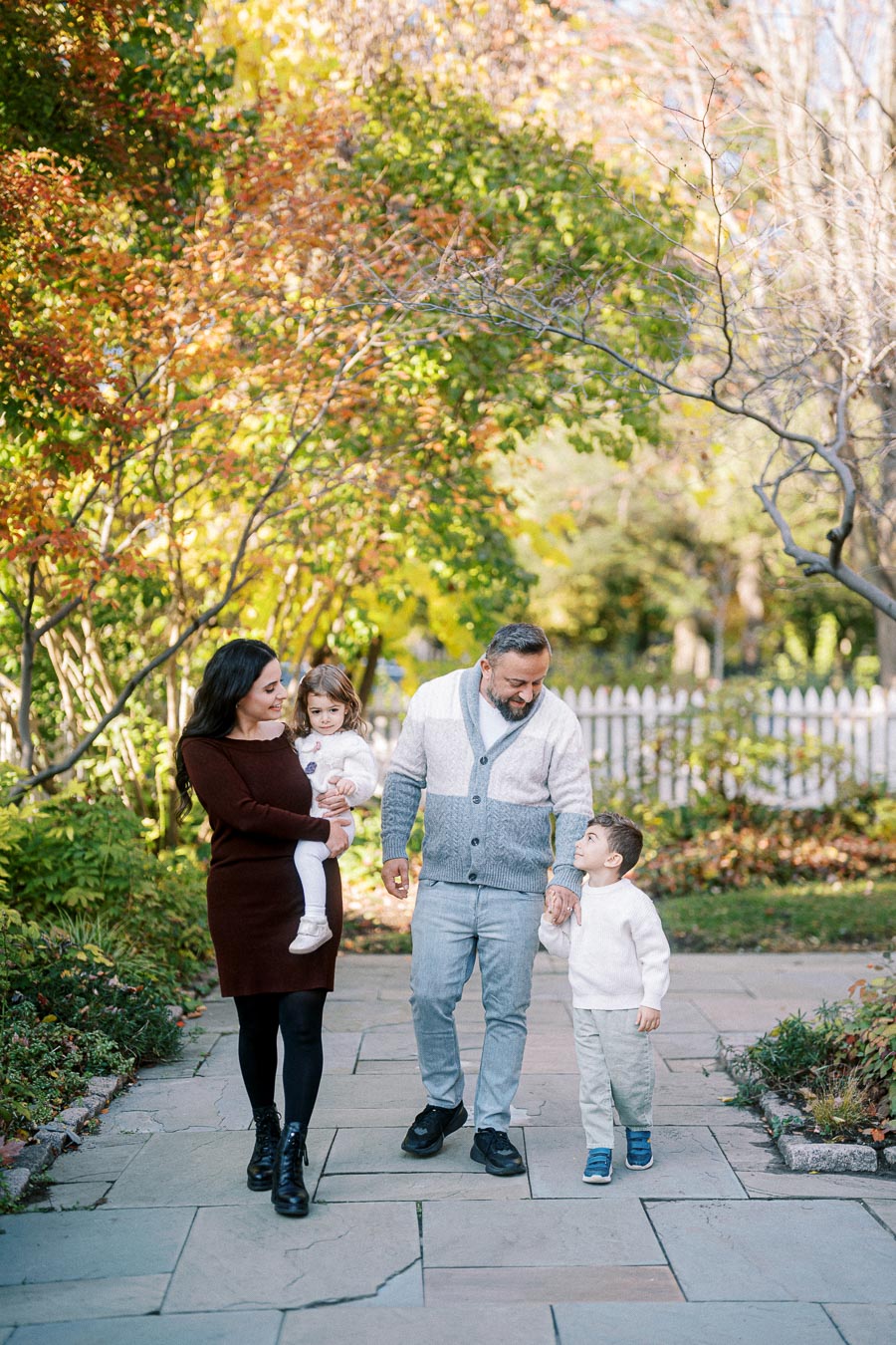 A family of four enjoying a walk in a picturesque park during autumn, with colorful foliage and a white picket fence in the background.