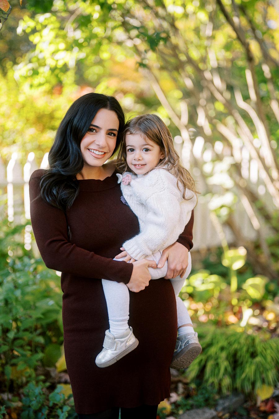 A woman in a brown sweater holding a young child wearing a white sweater and silver shoes, smiling outdoors with green trees and a white fence in the background.