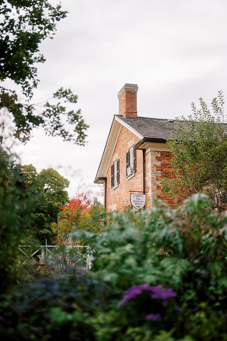 Historic brick house surrounded by lush greenery and colorful autumn foliage under a cloudy sky.
