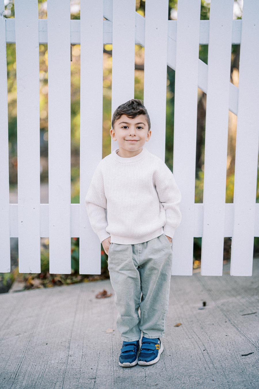 A young boy stands in front of a white picket fence, wearing a white sweater, light gray pants, and blue shoes, with hands in pockets and a slight smile, against a blurred green background.