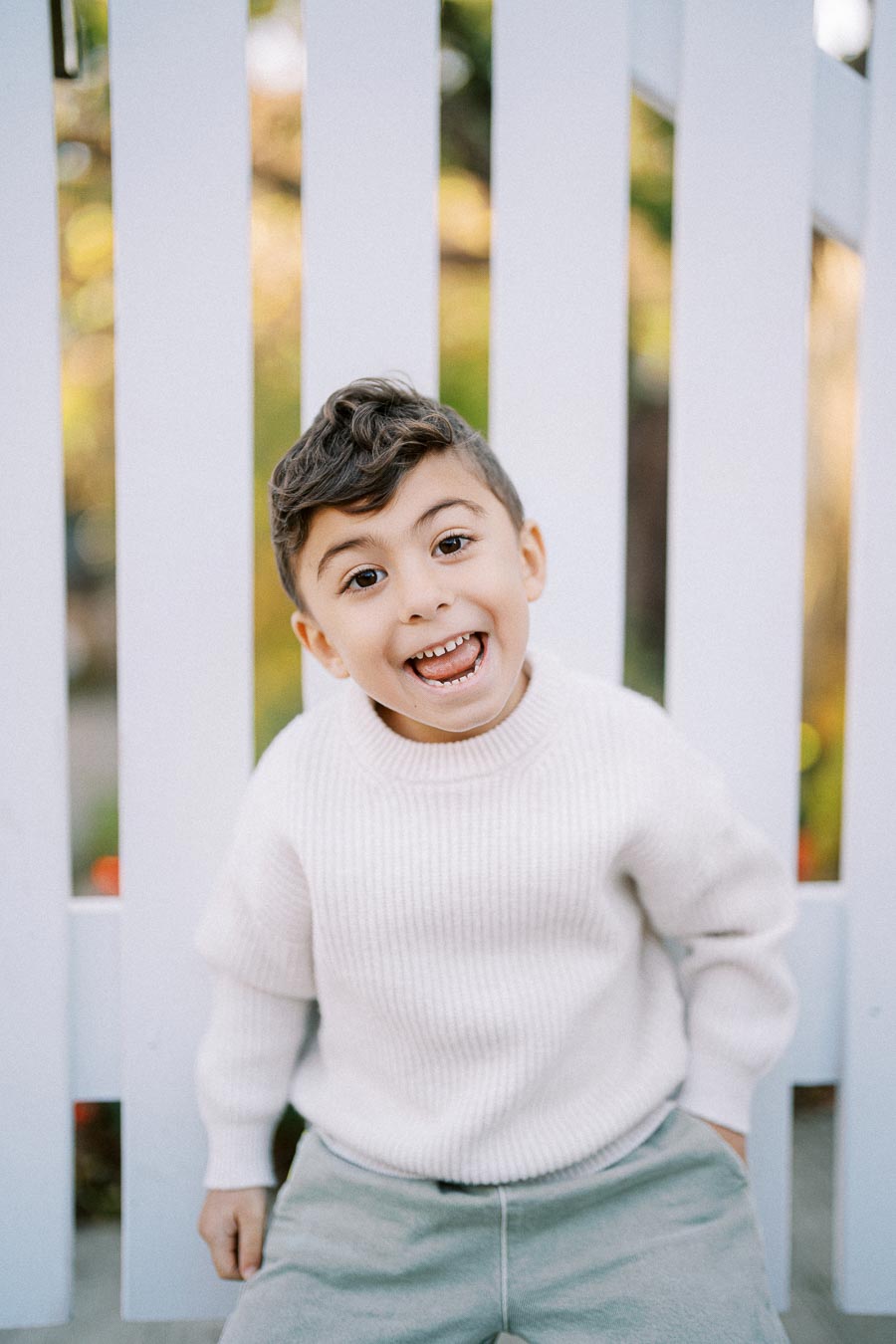 A cheerful young child wearing a cream sweater and light grey pants poses in front of a white picket fence with a joyful expression.