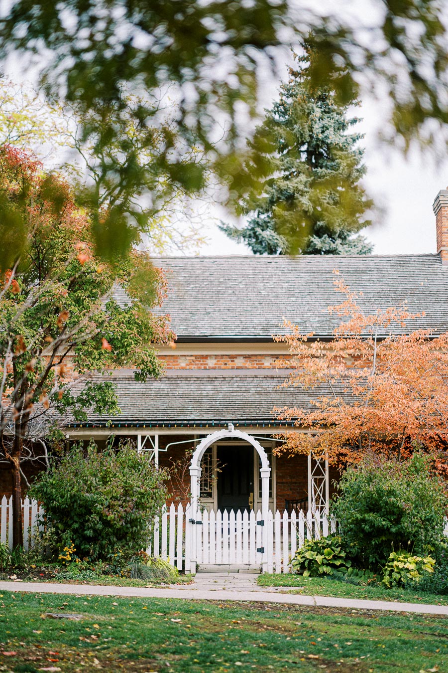 Charming brick house with white picket fence and archway, surrounded by lush greenery and autumn foliage, set against a peaceful suburban backdrop.