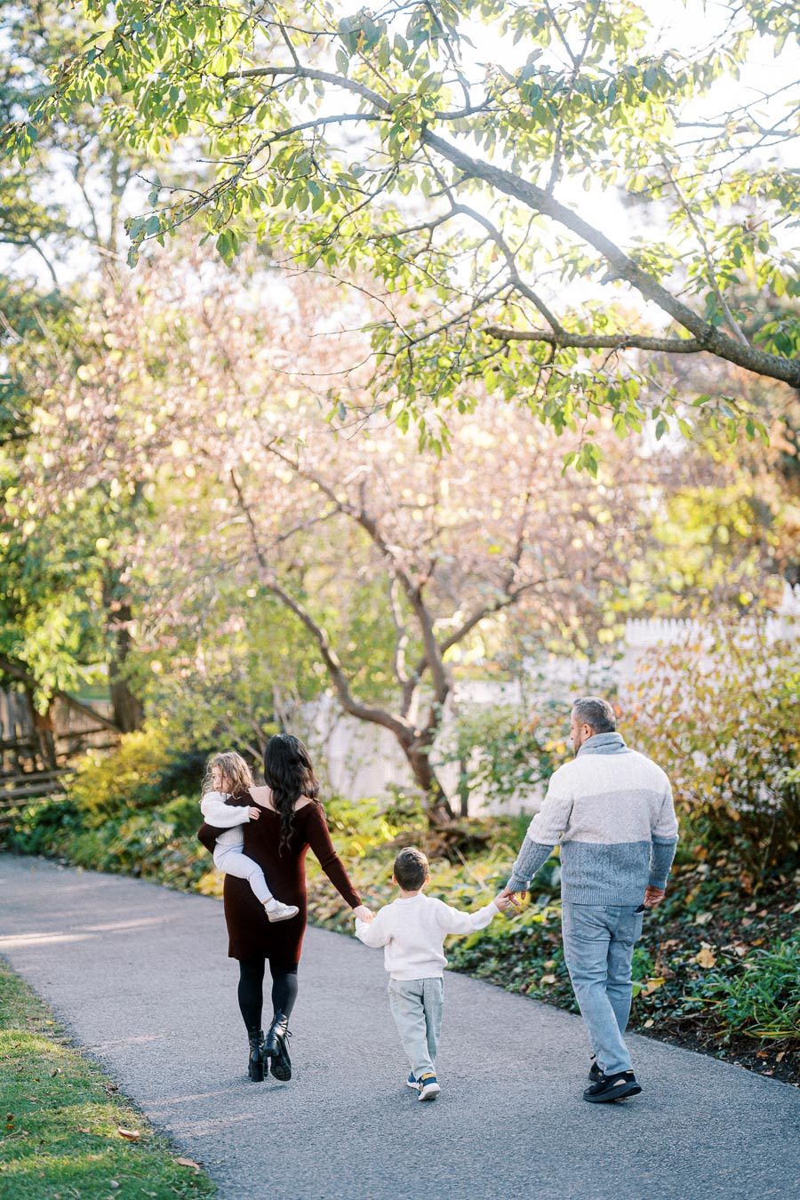 Family walking on a scenic park path surrounded by lush green trees during a sunny day.
