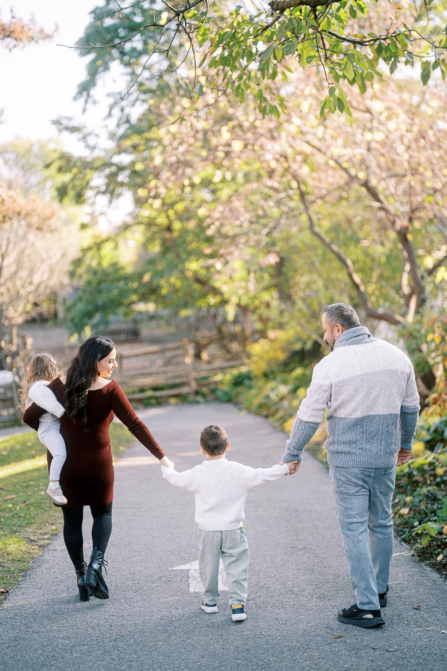 Family walking hand in hand down a tree-lined path during a sunny day in autumn, with lush green and golden foliage creating a serene and peaceful atmosphere.