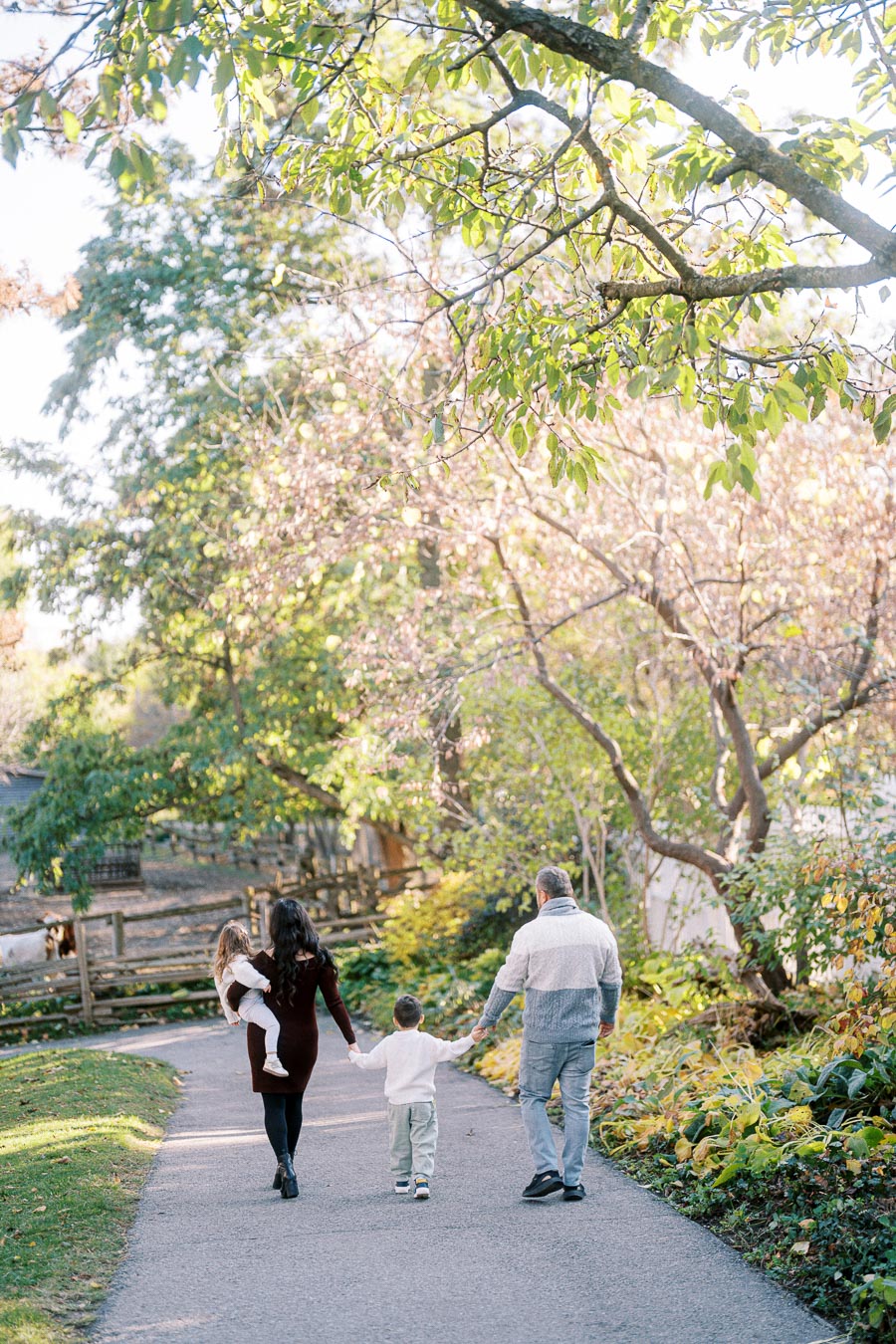 A family of four walking on a scenic path through a sunlit park, surrounded by lush green foliage and trees, capturing a serene outdoor moment in nature.