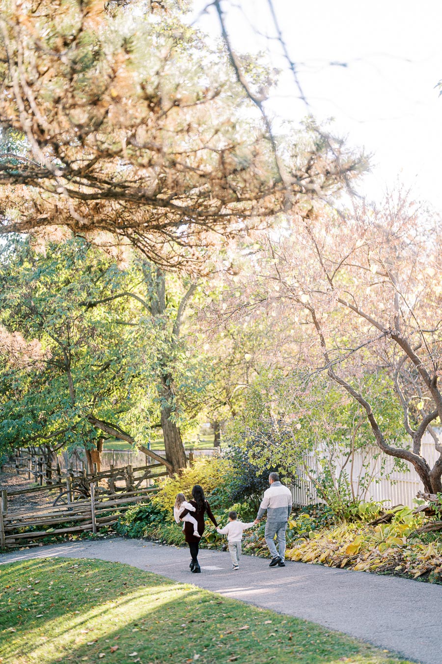 A family of four, including two children, walking along a tree-lined path in a serene park during autumn, with colorful foliage and sunlight filtering through the branches.