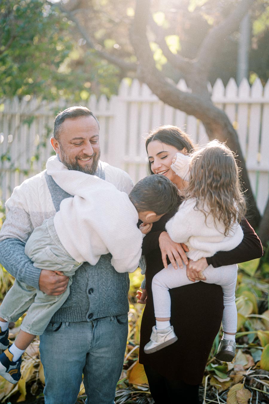 Happy family enjoying a sunny day in the garden, parents holding children, with a white picket fence and greenery in the background.