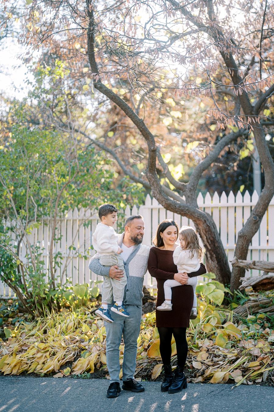 Family enjoying a sunny day in a lush garden, with two adults holding children, surrounded by greenery and a white picket fence.