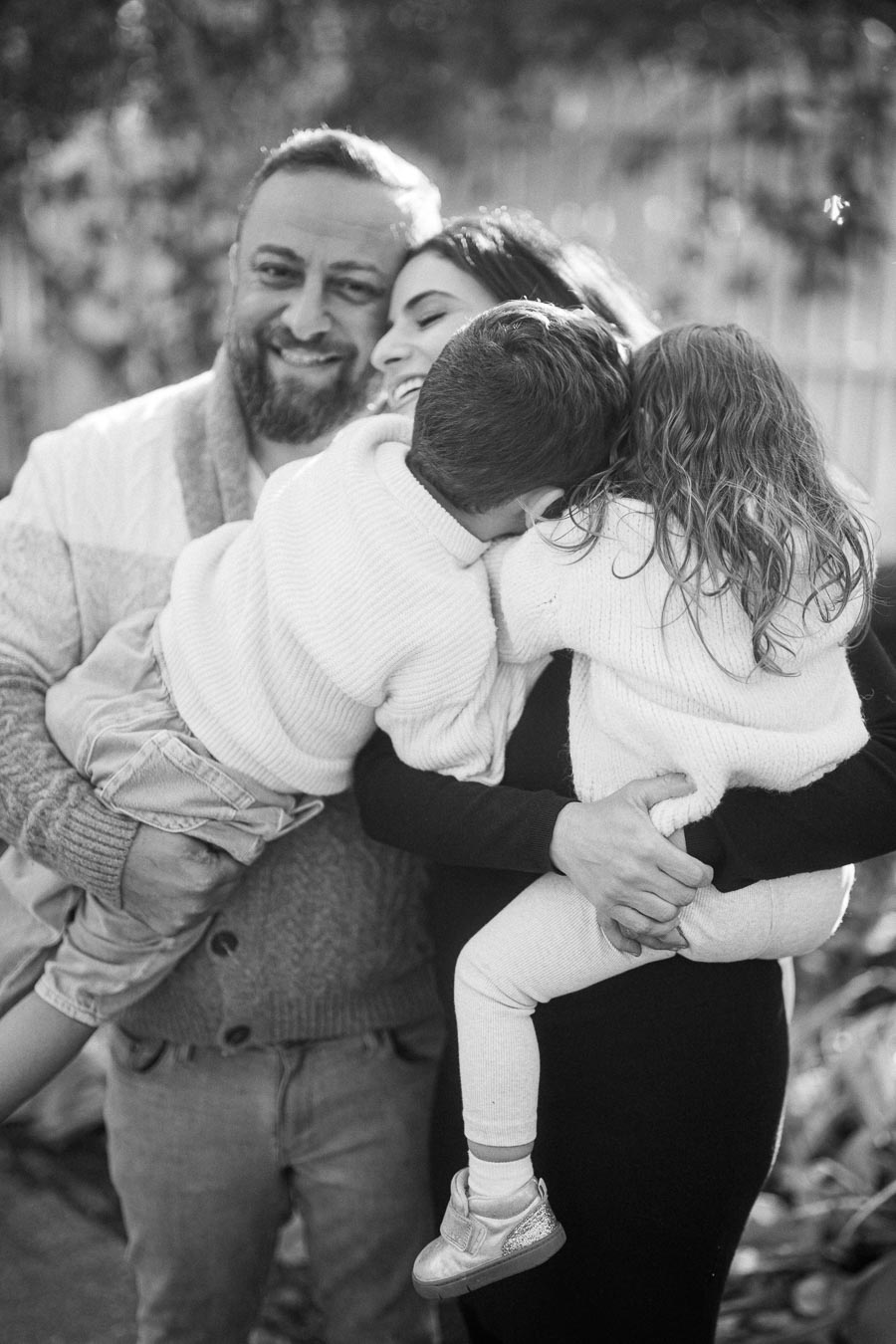 A loving family embracing outdoors, with parents holding their two young children closely, all wearing cozy sweaters in a black and white image.
