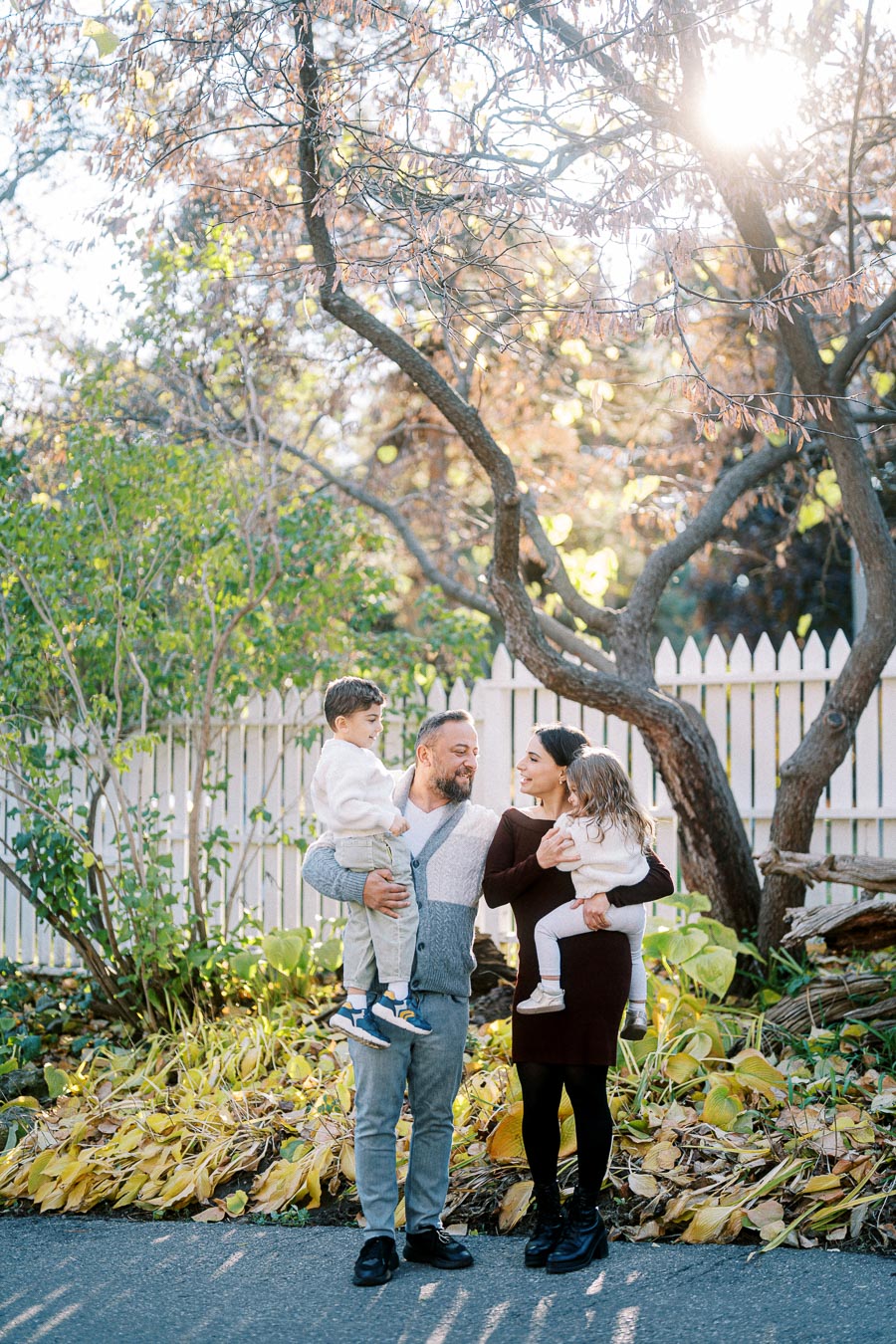 A family of four standing together outdoors in a garden with autumn foliage, smiling at each other under a tree with sunlight filtering through the branches.