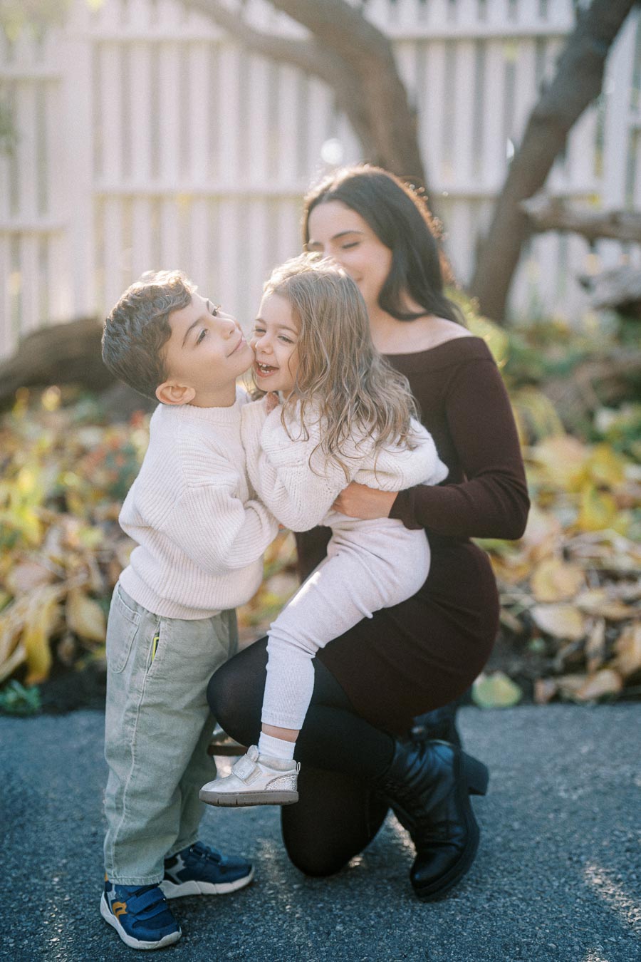 A mother kneeling on the ground embraces her young son and daughter, who are both smiling and wearing cozy white sweaters, in a sunny outdoor setting with autumn leaves and a wooden fence in the background.