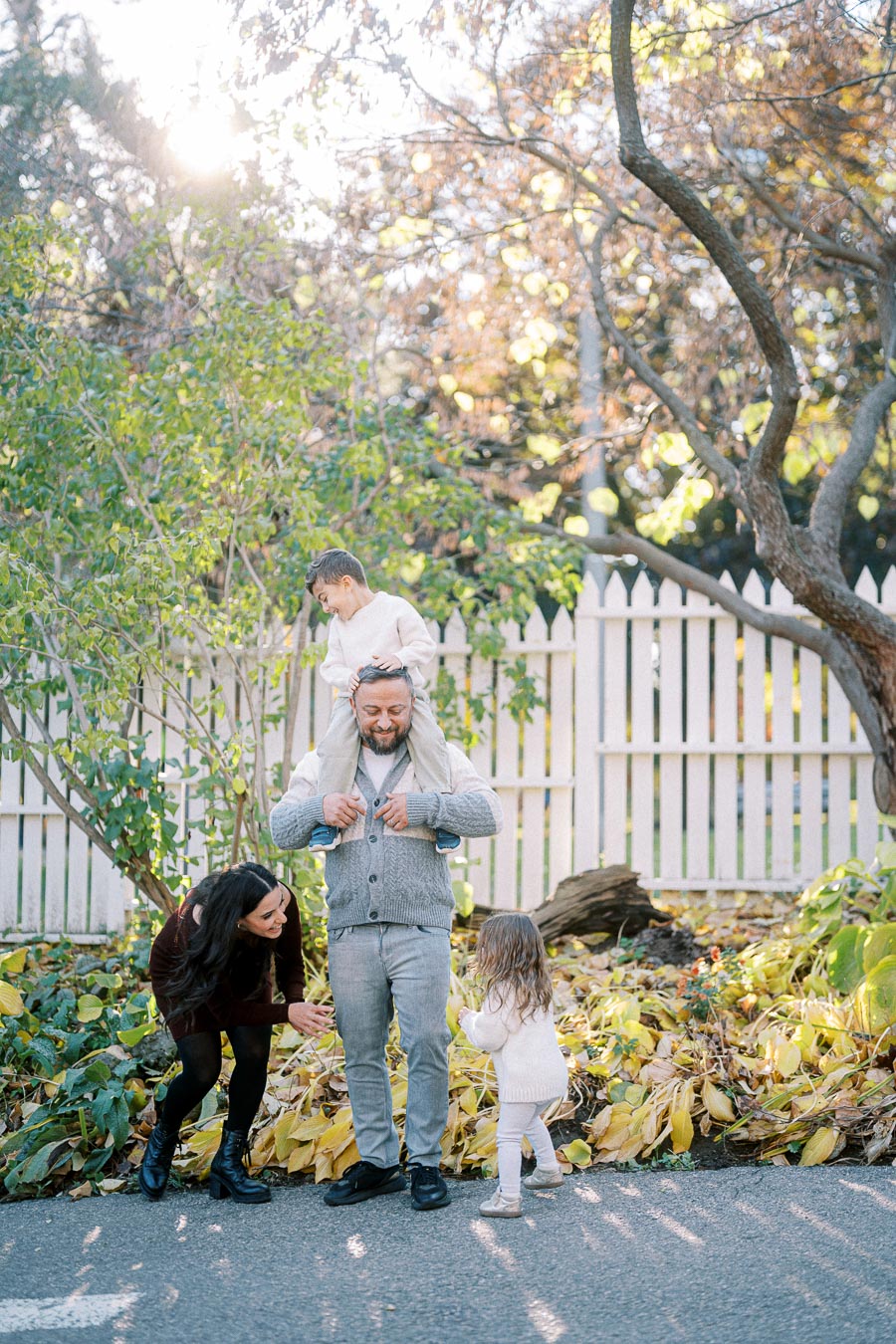A happy family enjoying a sunny day outdoors with a father giving his son a piggyback ride, a mother smiling at their daughter, and autumn leaves in the background near a white picket fence.