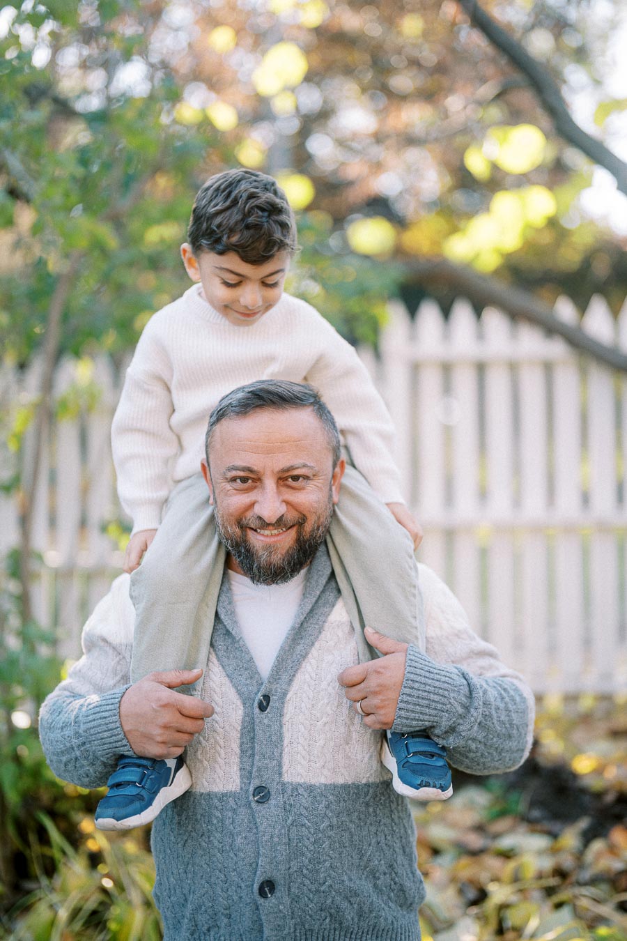 Father and son having fun outdoors, smiling as the child sits on his dad's shoulders, surrounded by a garden and wooden fence background.