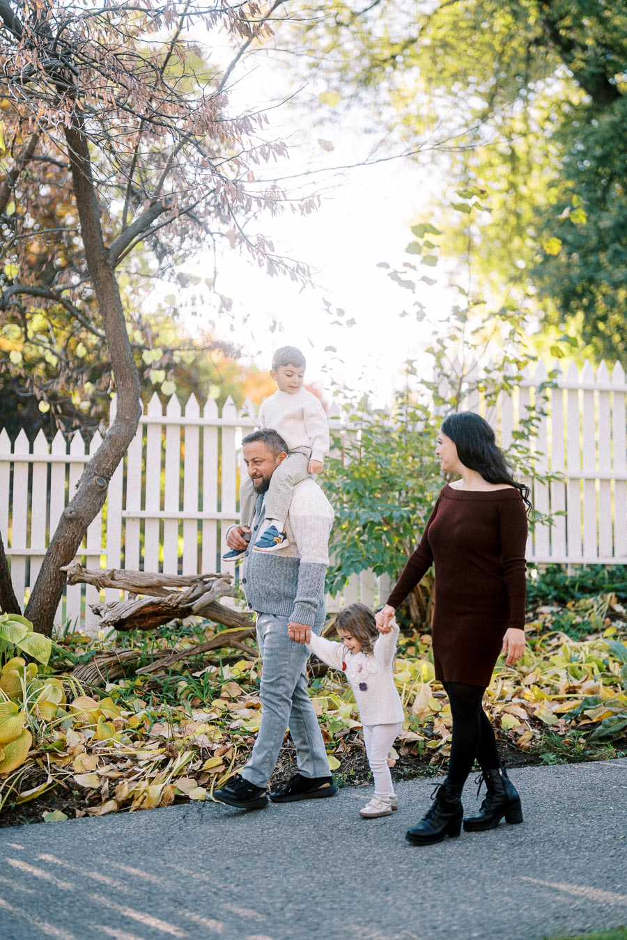 A family enjoying a walk in a park during autumn, with a person carrying a child on their shoulders and holding hands with another child, bordered by a white picket fence and colorful foliage.