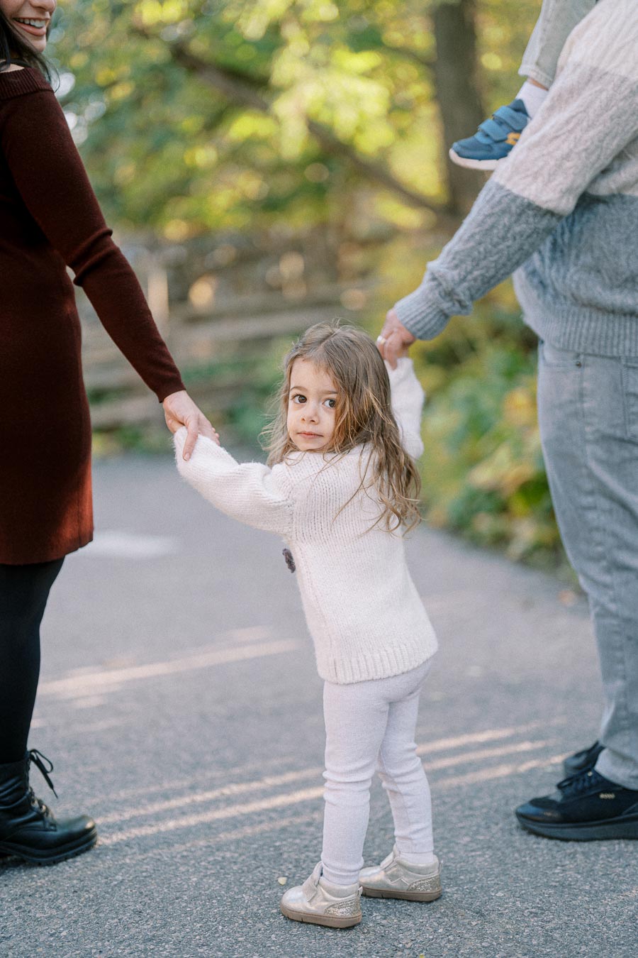 A young child in a white sweater and leggings holds hands with two adults while walking on a path in a lush park setting. The child looks back with a curious expression, surrounded by greenery and soft sunlight filtering through the trees.