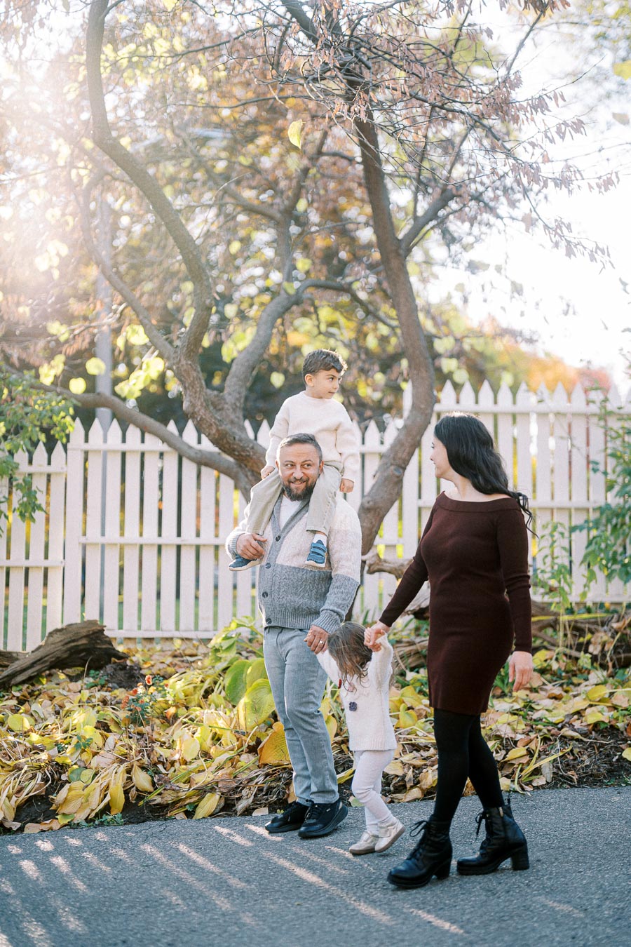 Happy family walking together in a park on a sunny autumn day, with children being carried and holding hands, surrounded by colorful fall leaves and a rustic white picket fence.
