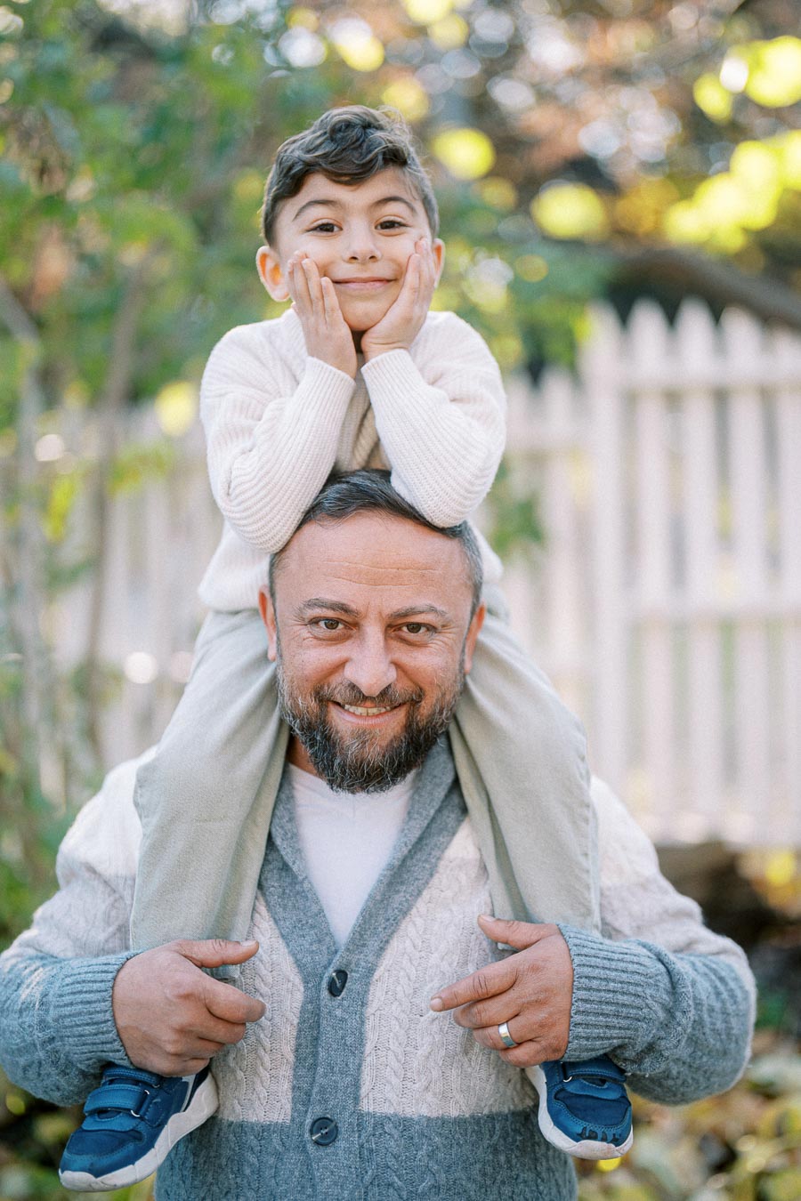 Father and son enjoying a playful moment outdoors, dad carrying boy on his shoulders against a blurred garden background.