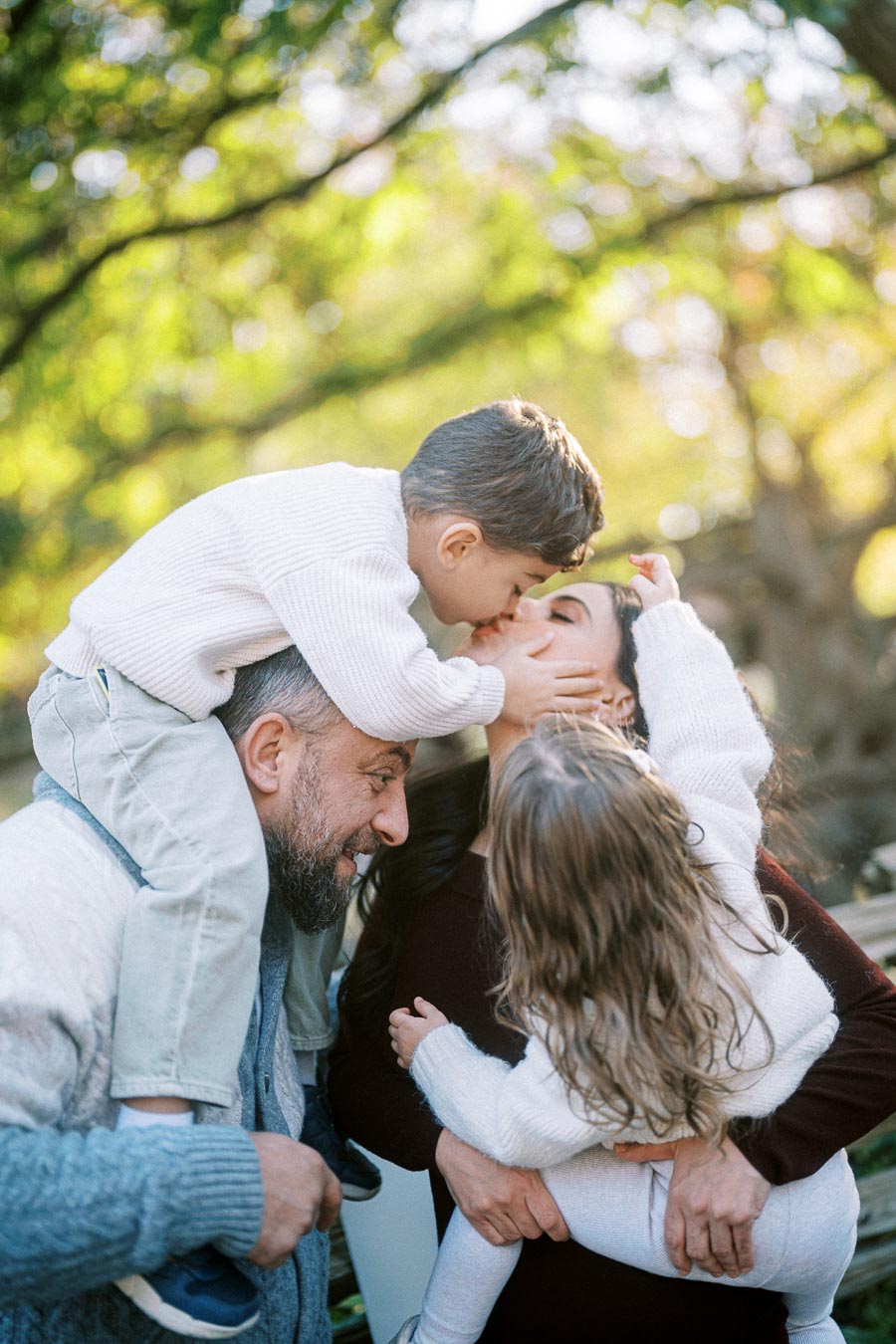 Happy family enjoying playful moments in a sunlit park, featuring a child on a man's shoulders kissing a woman's cheek while another child watches, surrounded by lush green foliage.