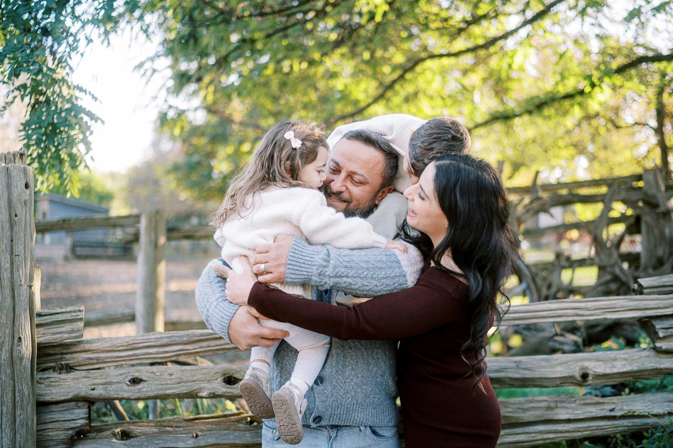 A family enjoying a loving group hug near a rustic wooden fence in a green, sunlit park setting.