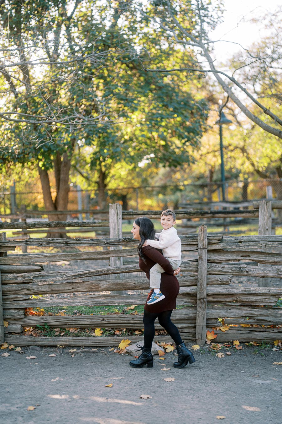 A woman gives a piggyback ride to a child in a scenic outdoor park with wooden fencing and trees in the background during a sunny day.
