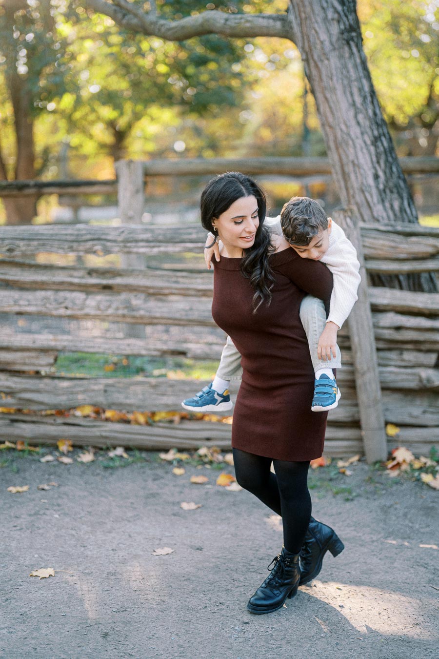 A woman in a maroon dress carries a young boy on her back while walking in a park during autumn, with a wooden fence and colorful leaves visible in the background.