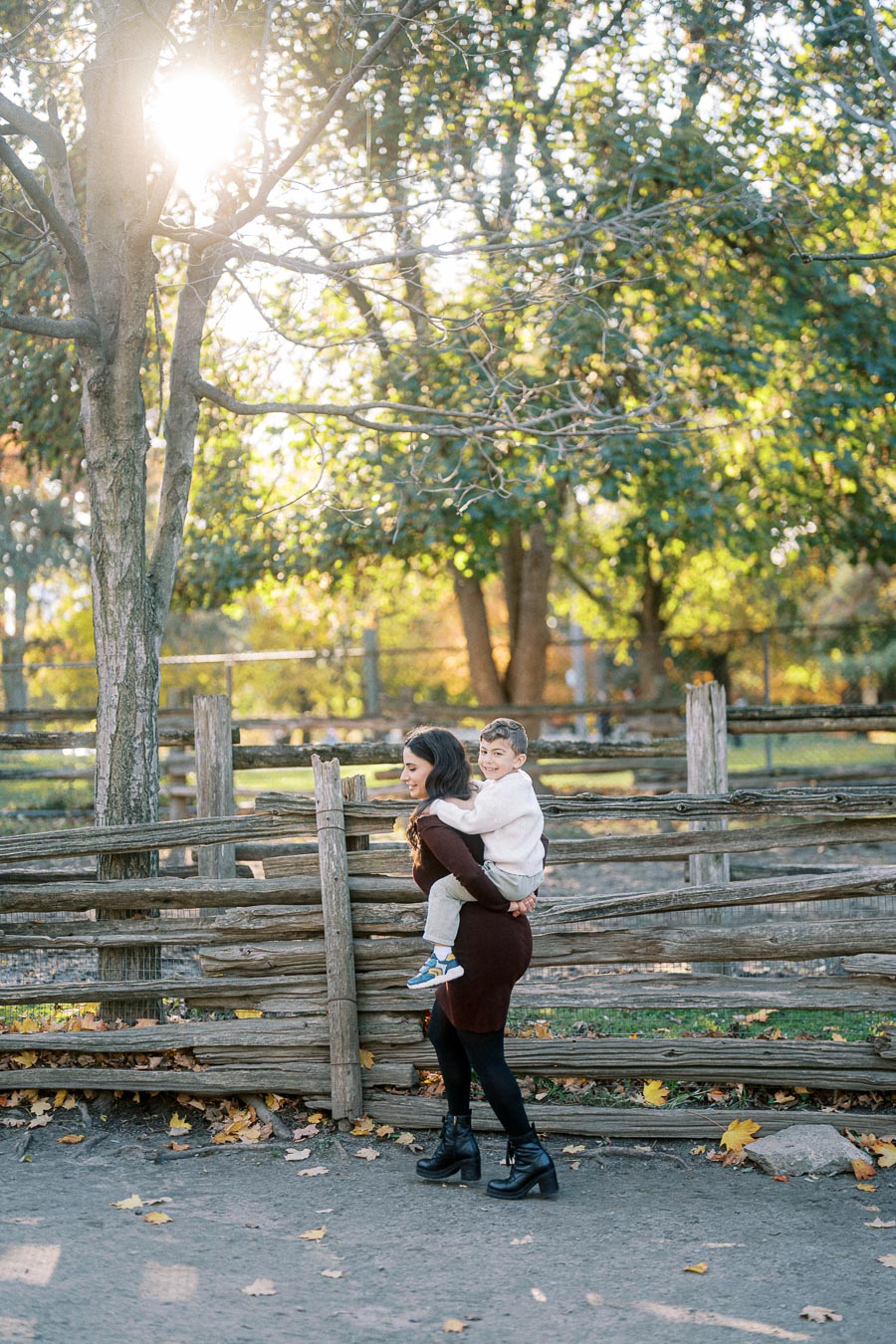 A woman giving a piggyback ride to a smiling child in a park, with sunlight filtering through the trees and a rustic wooden fence in the background.