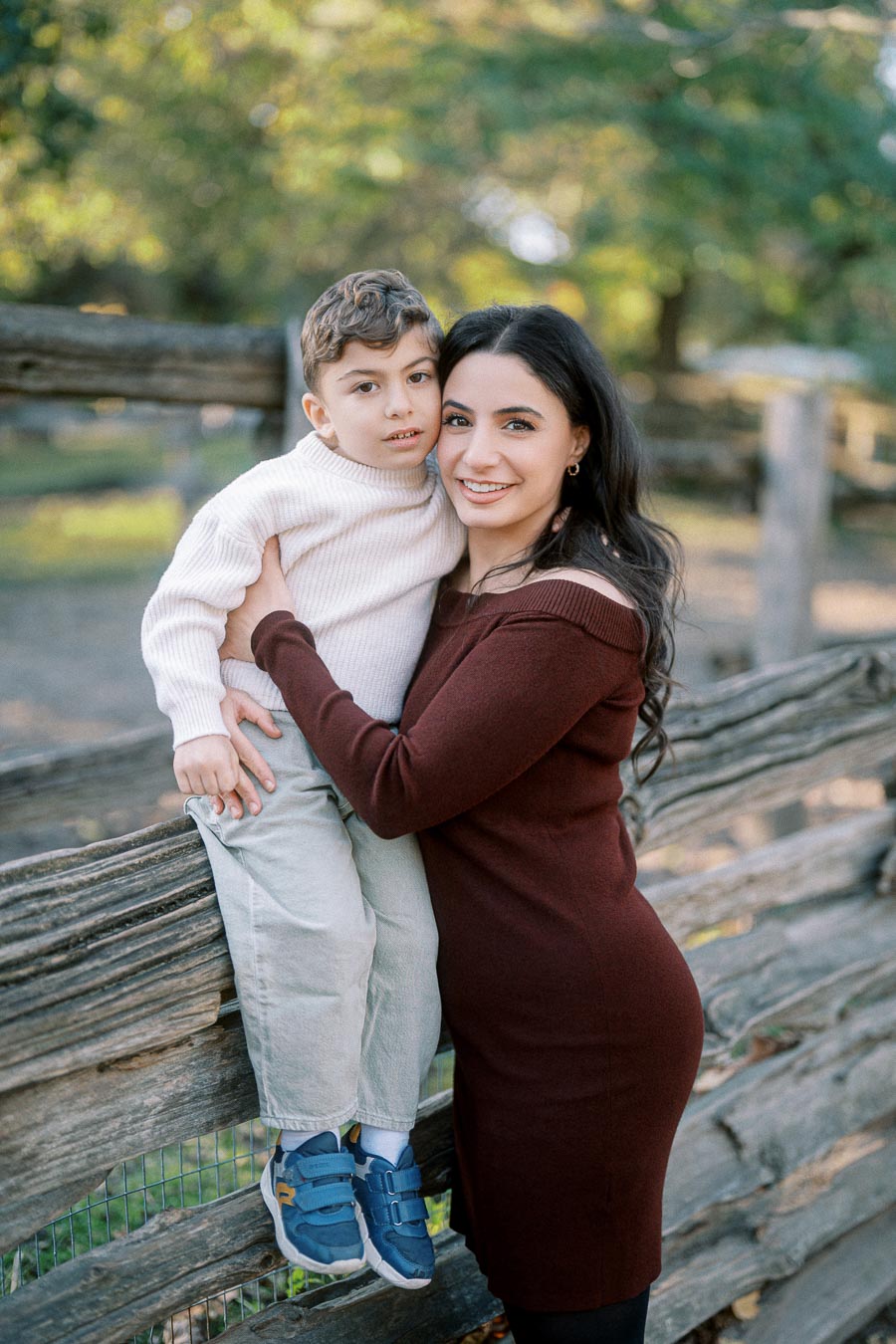 A mother holding her young son while leaning against a rustic wooden fence in a park during autumn, surrounded by trees and soft sunlight.