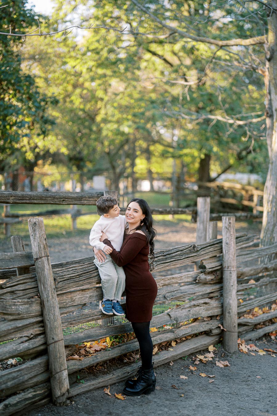 A mother and child enjoy a sunny day at the park, standing by a rustic wooden fence with lush green trees in the background. The mother, wearing a brown dress, is smiling while holding her young son, who is dressed in a white sweater and blue sneakers. The scene captures the warmth and joy of a family moment in nature.