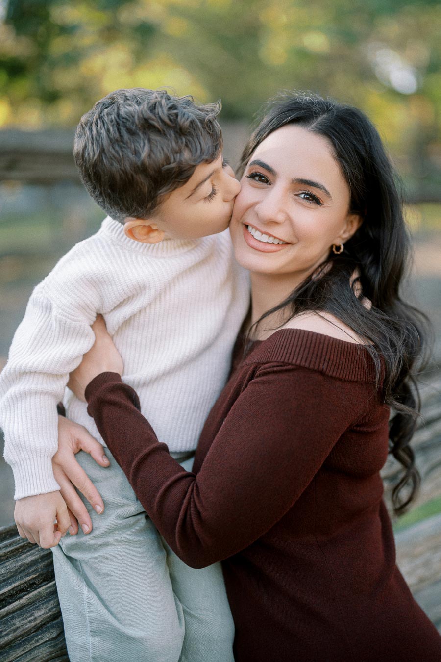 A joyful moment of a mother and her young son in an outdoor park setting, with the boy giving her a kiss on the cheek.