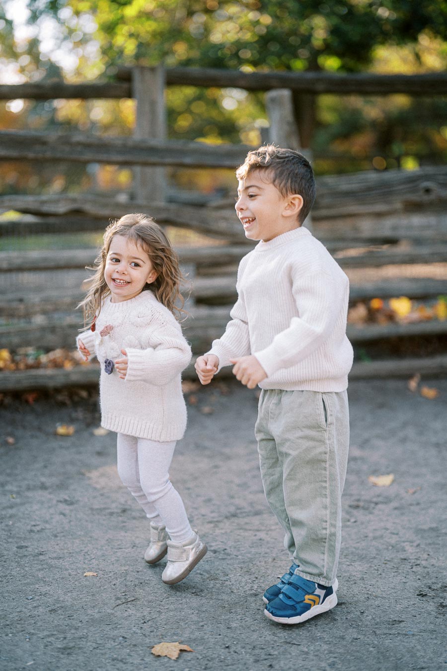 Two cheerful children playing outdoors, wearing cozy sweaters, with a wooden fence and autumn foliage in the background.