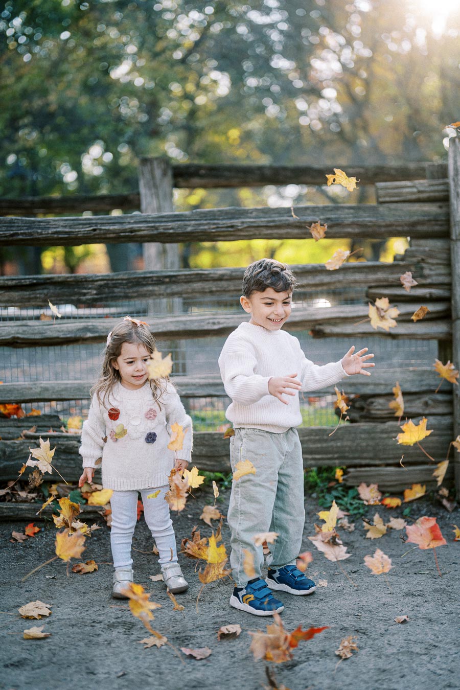 Two children joyfully playing with falling autumn leaves in a bright, sunny park with a wooden fence backdrop.