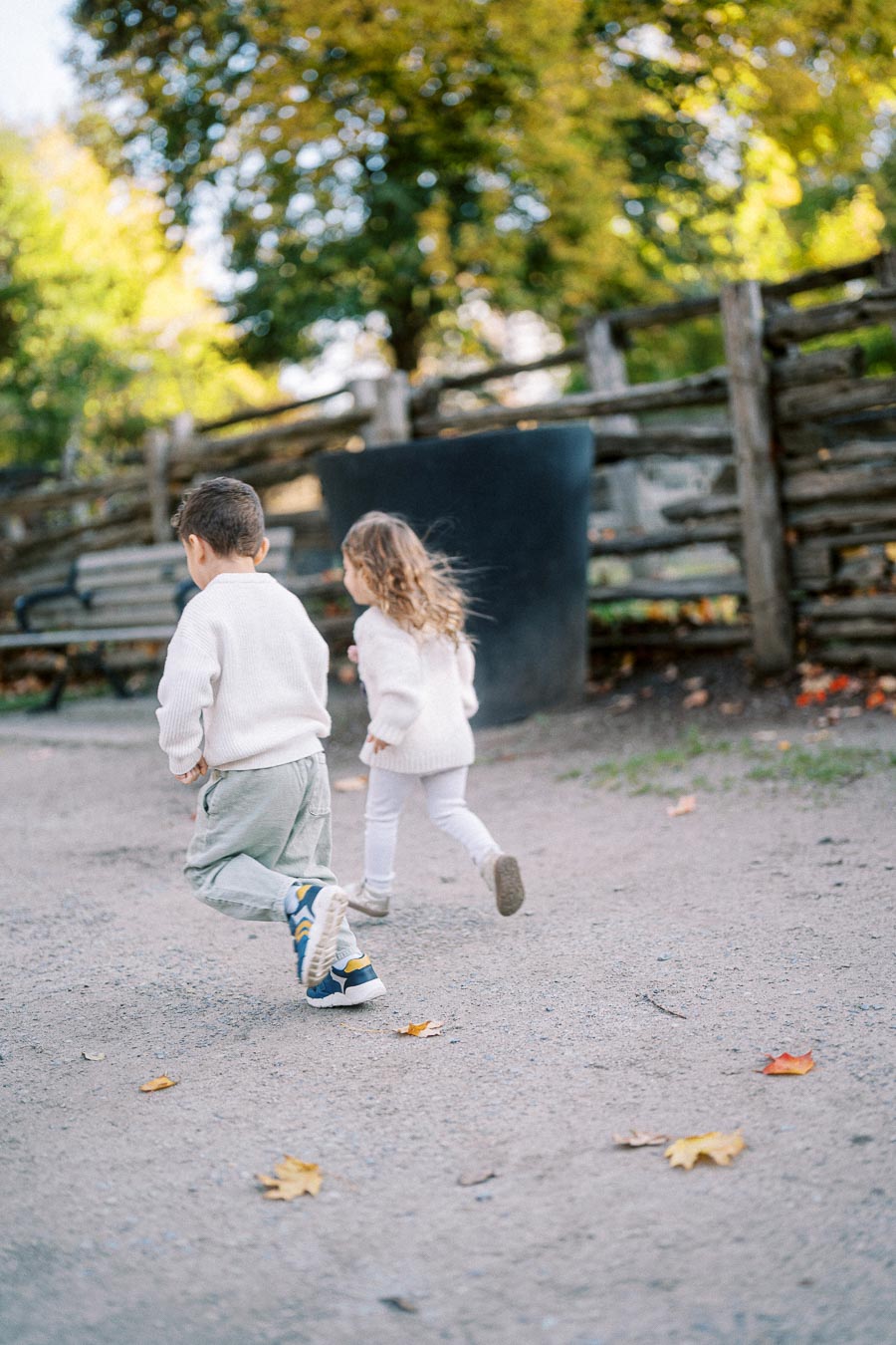 Two young children running joyfully on a park path with autumn leaves scattered on the ground, wearing cozy sweaters, surrounded by wooden fences and trees with fall foliage.