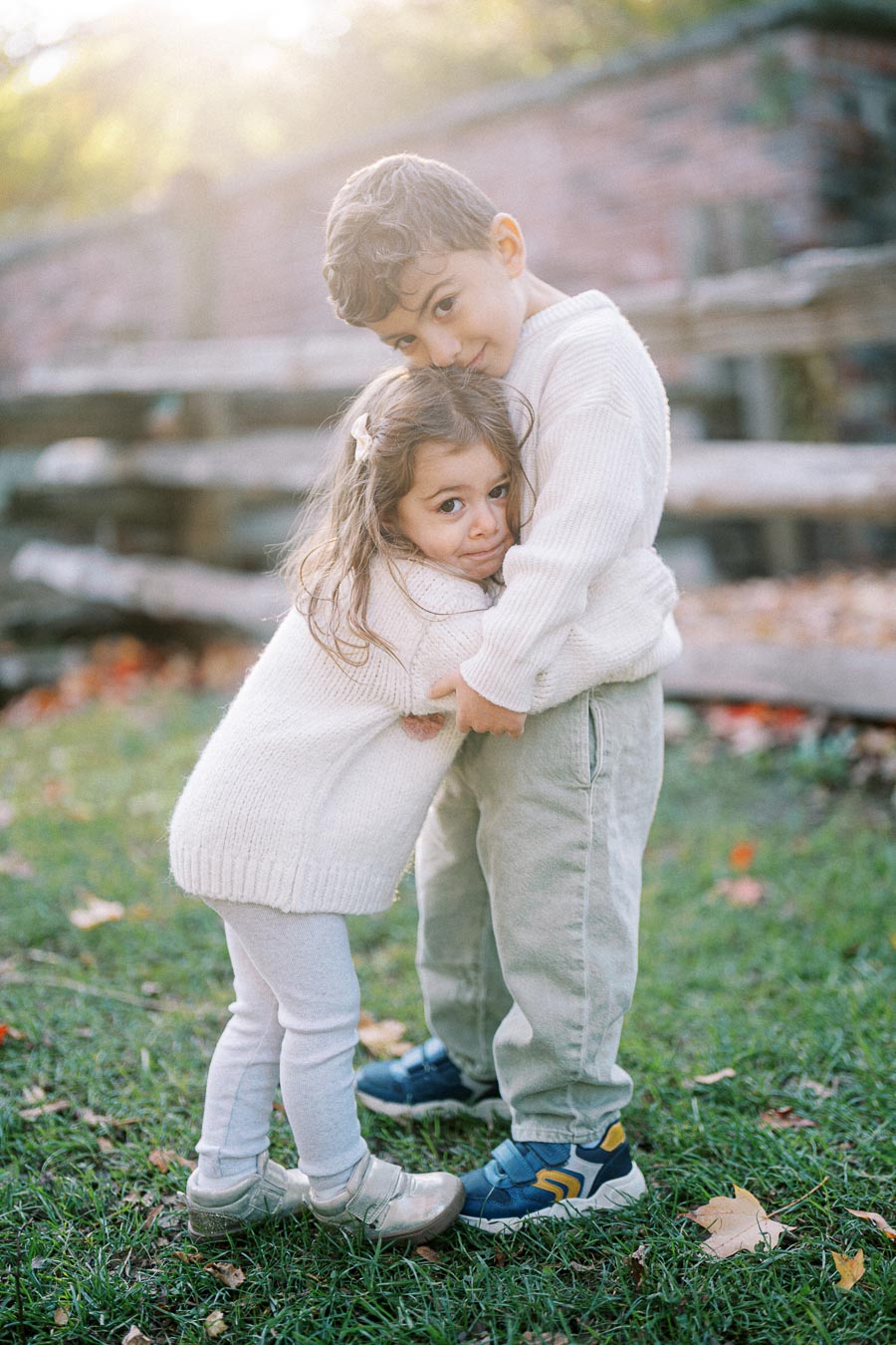 Two young children hugging outdoors in autumn, wearing cozy white sweaters, with fallen leaves and a wooden fence in the background.
