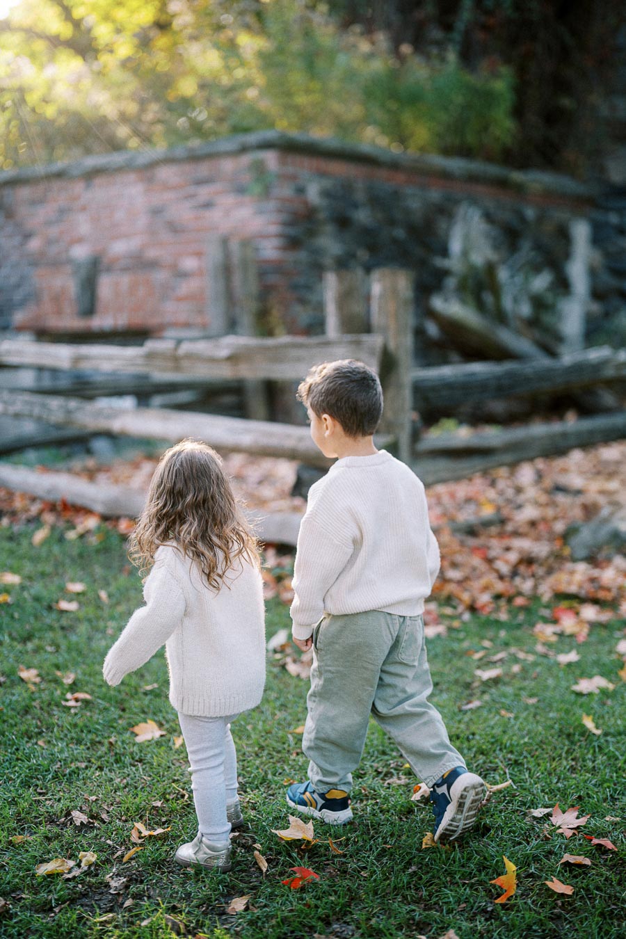 Two children wearing cozy sweaters walk through a grassy area covered with autumn leaves, with a rustic wooden fence and brick structure in the background, capturing the essence of a peaceful fall day.