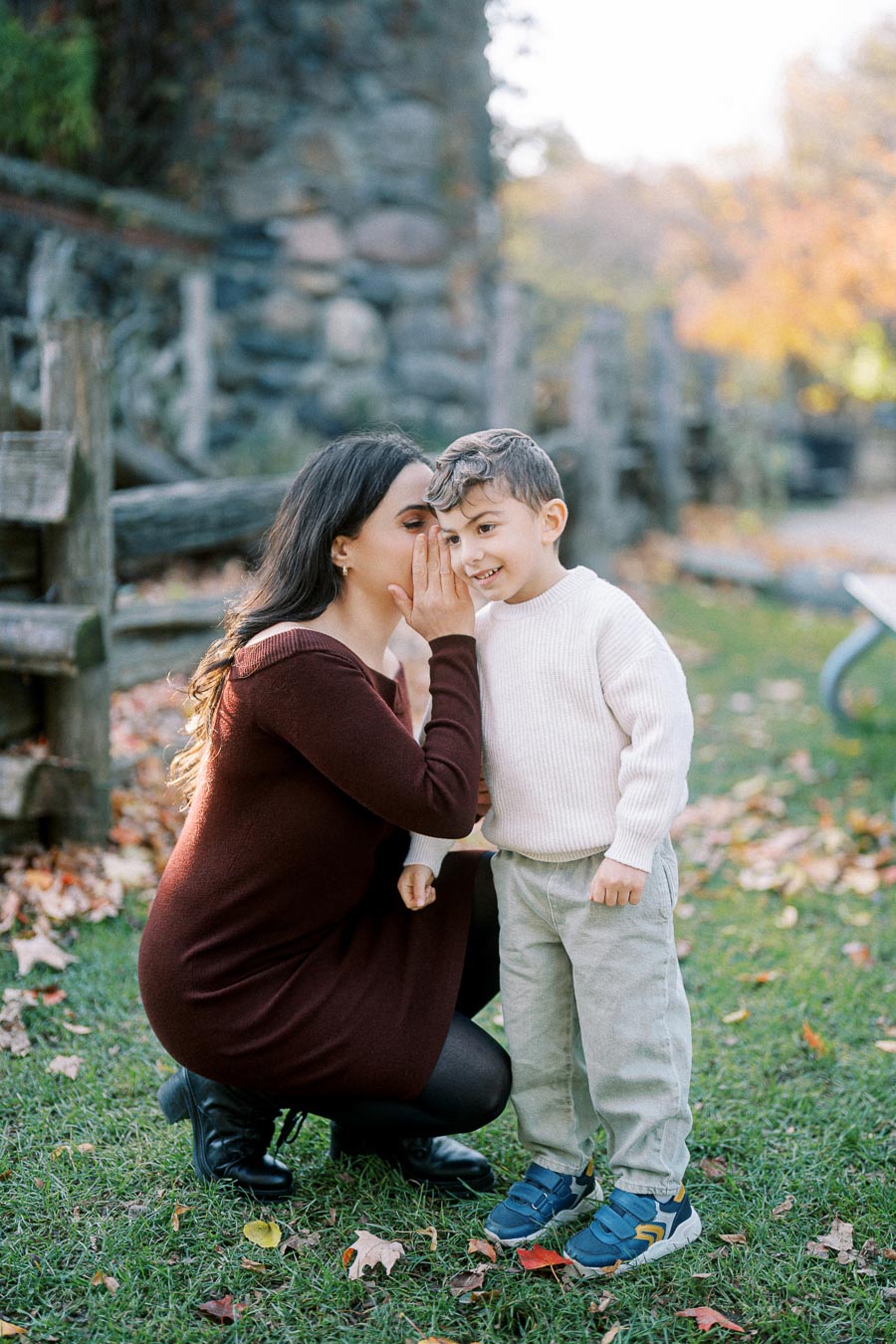 A woman in a maroon dress whispers to a smiling young boy in a white sweater and gray pants, outdoors on a grassy area with autumn leaves and a stone fence background.