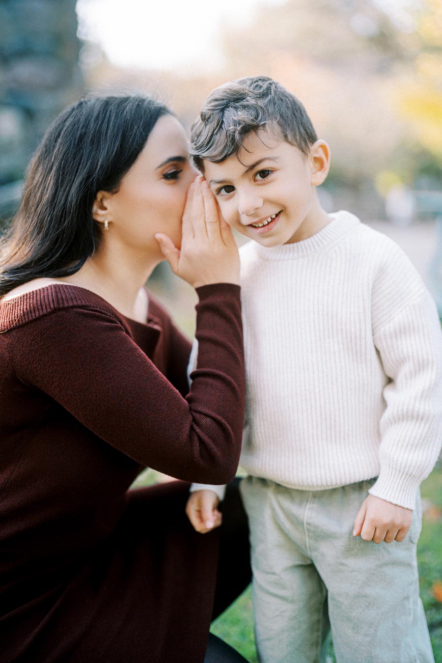 A woman whispers into a young boy's ear while he smiles outdoors, both dressed in autumn attire.
