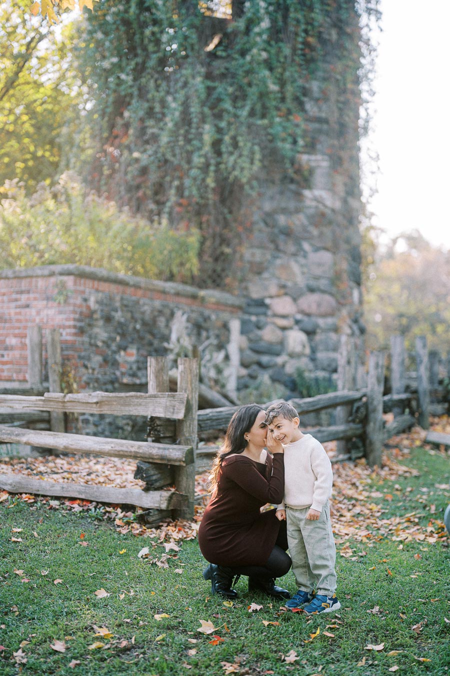A woman kneeling beside a young boy, sharing a secret in a sunlit park with autumn foliage, near a rustic wooden fence and ivy-covered stone wall.