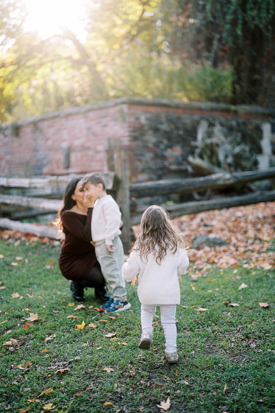 A mother embracing her young son while another child approaches, in a sunlit autumn park setting with fallen leaves and a rustic wooden fence.