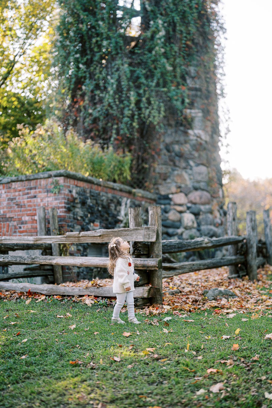 Young girl in white sweater standing near wooden fence, looking up at vine-covered stone tower amidst fallen autumn leaves.