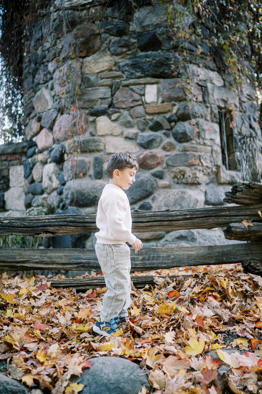 Young boy in a white sweater and jeans walking through colorful autumn leaves near a rustic stone building and wooden fence.